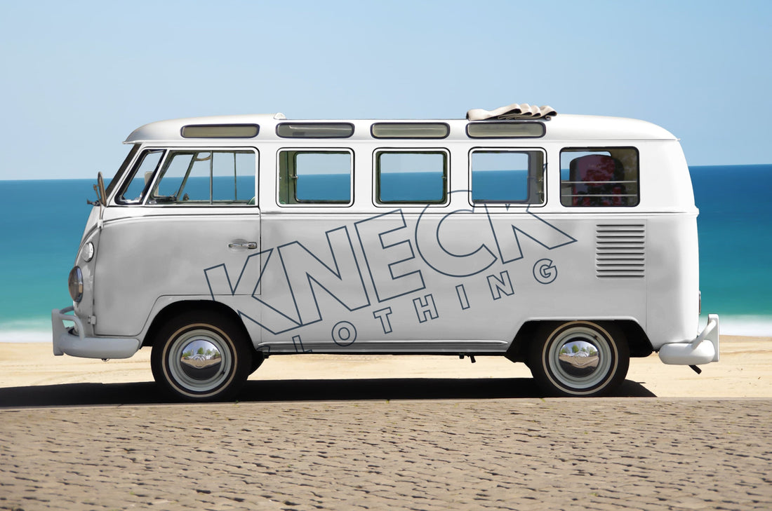 A white VW Camper van parked on a beach with the blue sea and blue sky in the background. The Camper has "KNECK Clothing" embossed diagonally on the side.